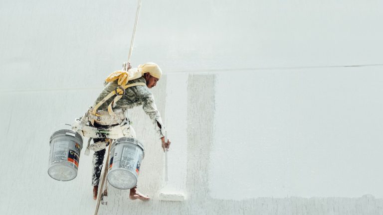 A painter suspended on ropes coating a large white exterior wall