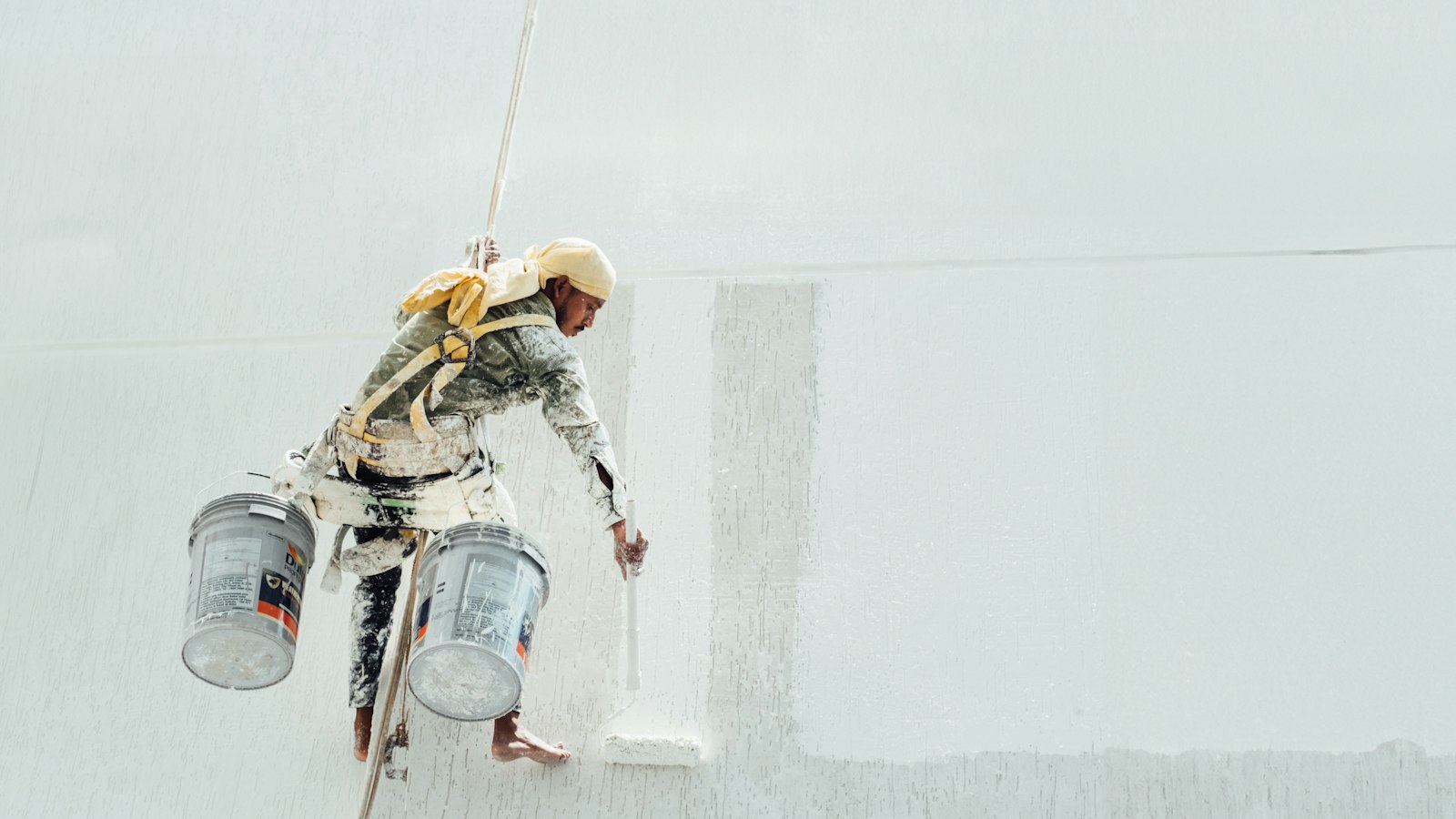 A painter suspended on ropes coating a large white exterior wall